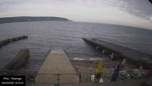 A view of the sea with a shoreline and two piers extending into the water. There is a ramp leading from the pebbly beach to the water's edge. On the right side, there are several boats, including a yellow kayak, a blue boat, and a red boat, all secured at the dock. The sky is overcast with soft clouds, and the cliffs are visible in the distance. The time and date are displayed at the bottom left corner.