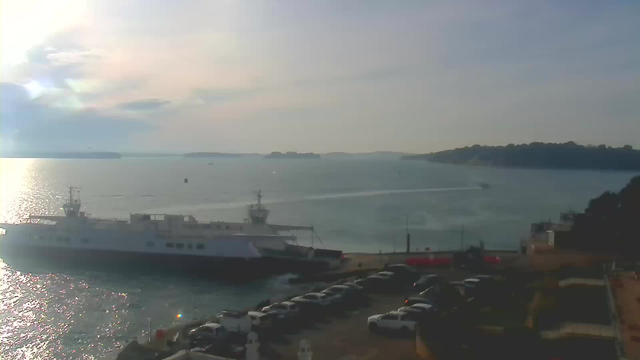 A large white ferry is docked at a waterfront, with parked cars visible in the foreground. The water appears calm with gentle ripples reflecting sunlight. In the background, a hazy shoreline stretches along the horizon, with further islands visible in the distance. The sky is partially cloudy, creating a serene atmosphere.