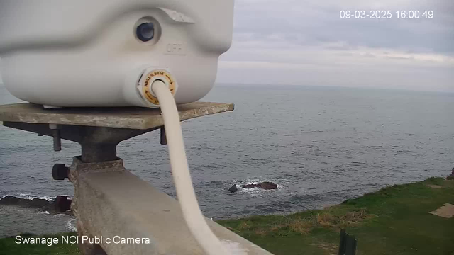 The image shows a close-up view of a white device attached to a metallic support, with a visible cable extending from it. In the background, the gray ocean stretches out under a cloudy sky, with a few rocky outcrops visible in the water. The shoreline features green grass and patches of land. A timestamp indicates the date and time as 09-03-2025 at 16:00:49.