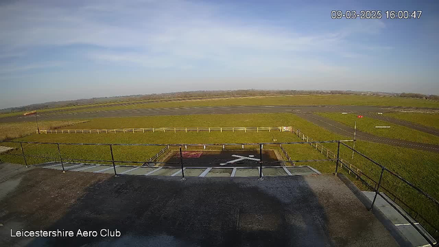 A wide view from a webcam at the Leicestershire Aero Club shows a grassy airfield. In the foreground, there is a flat rooftop with a railing. Below, a marked area for landing is visible with a white cross symbol. The airfield extends into the distance, bordered by a wooden fence. A runway can be seen on the right side, with a few small details like lights or markers. The sky is bright with some clouds, indicating clear weather.