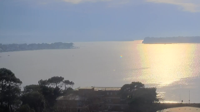 A panoramic view of a tranquil body of water under a sky with soft, diffused light. In the foreground, trees and a building are silhouetted against the water. The surface of the water reflects sunlight, creating shimmering patterns, while the shoreline features scattered boats. The distant scenery includes more trees and land extending into the water, suggesting a peaceful shoreline.