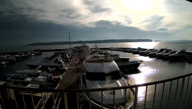 A view of a marina filled with various boats and yachts. The scene is cloudy with sunlight reflecting off the water, creating a shimmering effect. In the background, there is a distant landmass visible across the water. The foreground shows a wooden dock with boats moored alongside it.