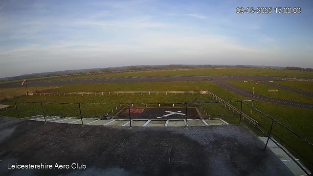 A view from a webcam at Leicestershire Aero Club shows a wide-open grassy area with an airstrip. In the foreground, there is a flat concrete structure with a railing and markings indicating a helipad, featuring a red and white design. The background displays a vast green landscape under a clear blue sky, with a faint outline of distant hills.