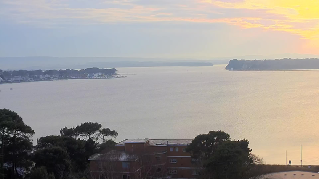 A panoramic view of a calm body of water at sunset. In the foreground, there are trees and a low-rise building with a flat roof. The water reflects soft pastel colors from the sky, ranging from light orange to pale blue. In the distance, a shoreline with houses and greenery is visible, and the horizon features rolling hills or additional land. Light clouds are dispersed across the sky, creating a serene atmosphere.