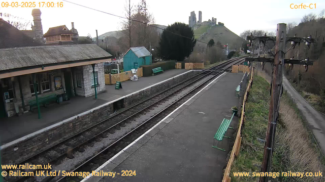 A view of Corfe Castle railway station featuring a gravel platform with green benches. In the background, there are tracks leading off into the distance. Beyond the tracks, a hillside is visible, with the ruins of Corfe Castle perched at the top. The scene is partly cloudy with a muted, early evening light. Alongside the platform, there is a wooden fence and a small green shed. A sign reading "WAY OUT" is visible on the platform.