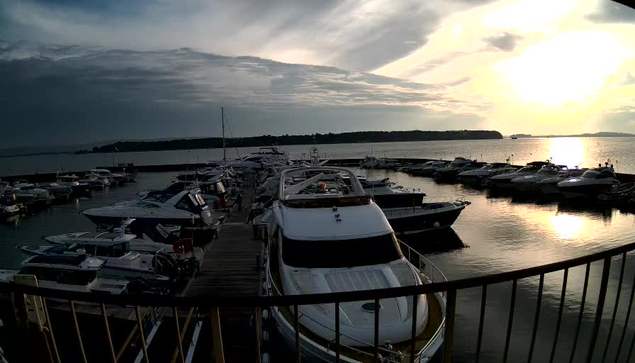 A marina scene at sunset, featuring several boats docked in calm water. The foreground includes a large white boat with a streamlined shape. Surrounding boats are mostly white and blue, some with visible cabins and sails. In the background, the sky is partly cloudy, with the sun glowing on the horizon, casting a reflection on the water. There are distant hills and trees lining the shore. A railing is visible in the lower part of the image, framing the scene.