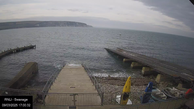 A view of a seaside location with calm waters under a cloudy sky. In the foreground, there are wide steps leading down to the water, bordered by a metal railing. A rocky shoreline can be seen, with waves gently lapping at the edge. To the right, there are two boats stored on the pebbled beach, one yellow and the other blue, along with visible sections of the beach. In the background, a long pier stretches out into the water, while cliffs rise in the distance.