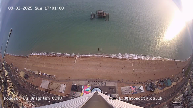 Aerial view of a sandy beach with a pier extending into the ocean. The beach is dotted with people enjoying the sun, and small waves are lapping at the shore. In the distance, there is a partially submerged structure in the water. Below, a promenade features colorful stalls and buildings. The scene is bright, reflecting a sunny day, with a slight shimmer on the water's surface. The date and time are displayed at the top of the image.