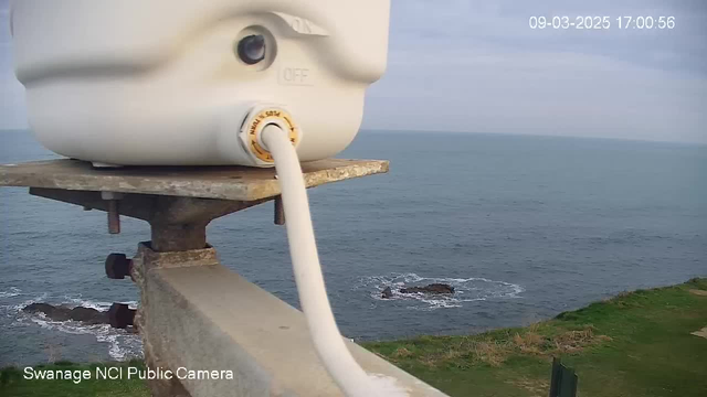 A close-up view of a white waterproof container, mounted on a metal platform, with a hose extending from it. The background features a calm sea, with a rocky shoreline and green grass visible in the foreground. The sky is partly cloudy, and the scene is taken from a public webcam.