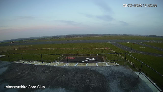 A panoramic view of a landscape from a webcam, showing a large, open field with a grassy area and a tarmac runway. In the foreground, there is a black area with a white cross symbol, indicating a landing zone. There is a fence surrounding part of the field and some trees in the distance. The sky above is mostly clear with soft clouds, and the lighting suggests it is late afternoon. A wind direction indicator is visible near the left side of the frame. The scene is calm and quiet, typical of an aerodrome setting.