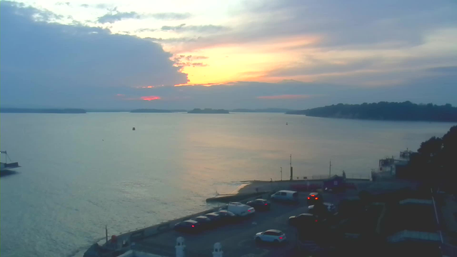 A tranquil evening scene featuring a calm body of water reflecting soft hues of orange, pink, and gray from the sunset above. The horizon is lined with distant islands and the shoreline includes a small dock with boats. In the foreground, there are several parked cars and a portion of the waterfront walkway, while low greenery and structures are visible along the edge of the image. The sky is partly cloudy, enhancing the serene atmosphere.