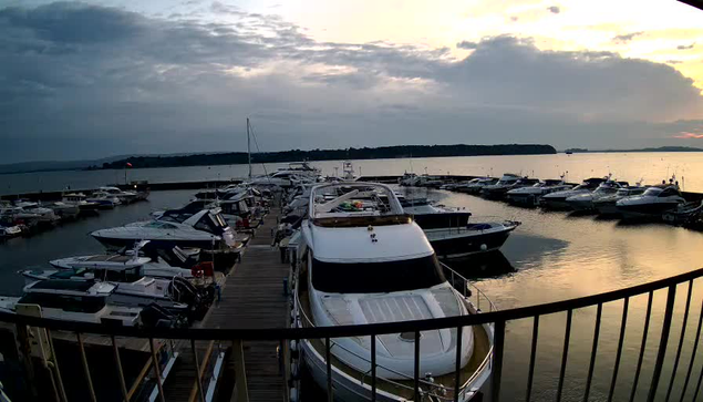 A marina scene at sunset, featuring several boats moored in calm waters. The foreground has a large white boat with a shiny surface, and wooden docks extending alongside. Various other boats of different sizes and colors are visible, with some distant land in the background under a cloudy sky. The sun is setting, casting a warm glow over the water, creating soft reflections and a serene atmosphere.