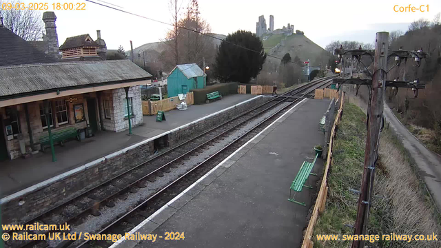 A railway station platform is depicted with several green benches situated along the edge. To the left, there is a stone building with a sloped roof, featuring large windows and a sign indicating "Corfe Castle". Behind the station, a hillside rises, topped with the ruins of Corfe Castle, surrounded by trees and greenery. Two sets of railway tracks run parallel across the foreground, leading off into the distance. A wooden fence and a small blue building are visible to the left, along with an entrance sign that reads "WAY OUT". The image captures the scene in late afternoon light.