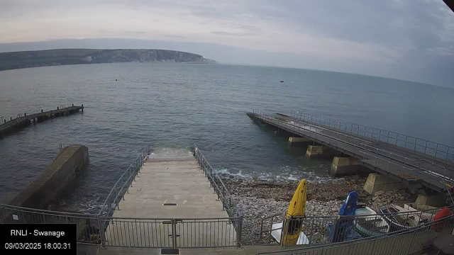 A view of a calm sea with gentle waves, shown from a concrete walkway leading down to the water. On the left, there is an incomplete stone pier extending into the water. To the right, a wider wooden pier also stretches into the sea. Along the shoreline, there are small yellow and blue kayaks and scattered stones. In the background, a hillside with greenery and cliffs rises above the water, under a cloudy sky.