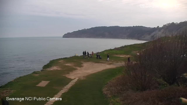 A coastal scene featuring a calm sea under a cloudy sky. In the foreground, there is green grass and some sandy patches. A group of people, including individuals walking and standing, gather near the edge of the water. In the background, a rocky cliff rises above the coastline. The atmosphere is serene, with no visible signs of human structures nearby.