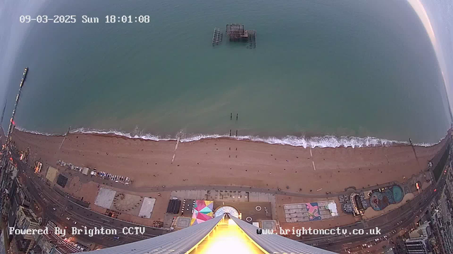 Aerial view of a beach with soft, sandy shore and gentle waves lapping at the edge. In the water, an old pier structure is visible, partially submerged. The beach is dotted with a few people walking along the shore. Colorful amusement park rides and structures can be seen on the beach, with various patterns and colors. The image is taken at dusk, with a warm glow from a nearby structure reflecting the last light of day. The timestamp at the top indicates the date and time of the capture.