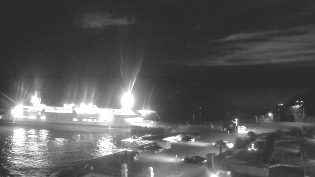 A nighttime image of a harbor scene. A large ferry is docked, illuminated by bright lights coming from the vessel and surrounding areas. The water reflects these lights, creating a shimmering effect. There are several cars parked nearby and a roadway leading up to the dock. In the background, distant lights on land suggest buildings and trees, set against a dark sky with wispy clouds.