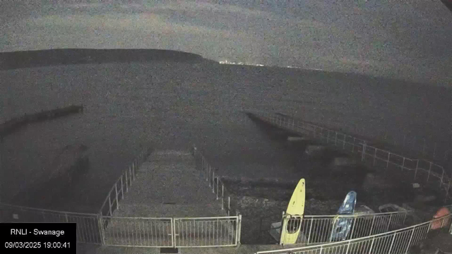 A dimly lit scene of a waterfront area. In the foreground, a walkway made of grey stones leads to a boat ramp with metal railings on either side. Two kayaks are parked on the right side, one yellow and one blue, partially visible. The water is dark and calm, reflecting soft lights in the distance. The sky is mostly cloudy with faint signs of dusk. The image has a timestamp in the lower left corner, indicating it was taken in Swanage.