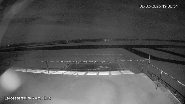 A low-light image of an airfield from a webcam at Leicestershire Aero Club. The foreground shows a flat surface with lines indicating the boundaries of a helipad, marked by a cross. A railing borders the edge of the platform, and the background features a dark sky with subtle outlines of the landscape and distant lights. The scene is mostly monochrome, given the low-light conditions. The timestamp indicates the image was captured at 19:00 on March 9, 2025.