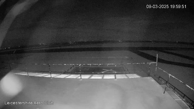 A dark scene captured from a webcam shows a flat area resembling an airport runway with markings in the center. The surrounding area is mostly dark, indicating it is nighttime, with faint lights visible in the distance. The image includes a railing in the foreground, and the timestamp in the corner reads 09-03-2025 at 19:59:51. The bottom left corner has the text "Leicestershire Aero Club."