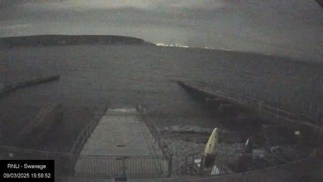 A dimly lit coastal scene featuring a pier extending into the water. The foreground shows a concrete platform with some equipment, including a yellow kayak and various objects. The ocean appears dark and slightly choppy, with a distant shoreline visible in the background, where faint lights indicate a nearby town. The sky is overcast, contributing to the overall muted atmosphere.