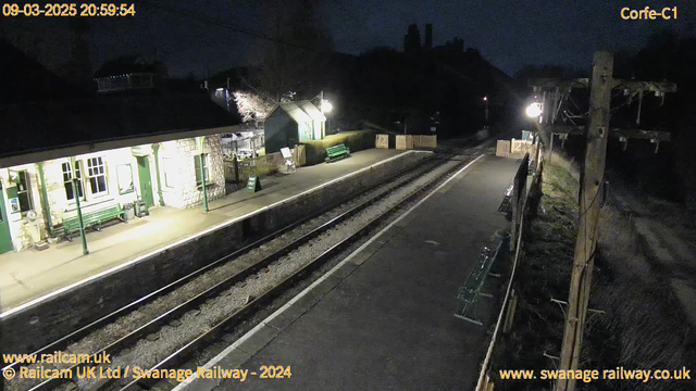 A dimly lit train station at night. The platform is empty with two railway tracks running parallel. On one side, there is a stone building with large windows and a green bench in front, along with a green sign that reads "WAY OUT." A wooden fence and some hedges are visible in the background, along with a few more benches and a light post illuminating the area. The surroundings are dark, with silhouettes of buildings and trees in the distance.