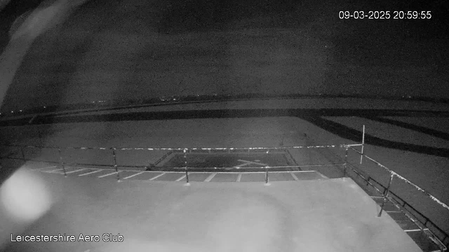 A webcam image showing a nighttime view of a tarmac at Leicestershire Aero Club. The scene is dark with limited visibility. A section of the tarmac is marked with a white "X" against a background of silhouetted outlines and faint lights in the distance. The image includes a railing in the foreground, and the timestamp of the capture is displayed in the upper right corner.