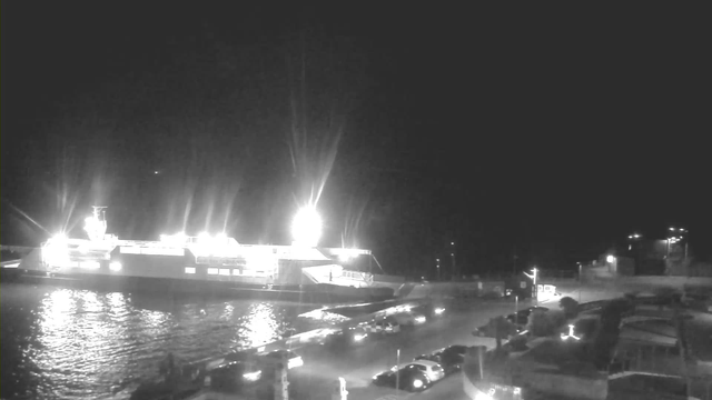 A nighttime webcam image shows a brightly lit ferry docked at a port. The ferry has strong lights illuminating its deck and surroundings. Calm water reflects the lights, creating a glowing effect. In the foreground, several parked cars are visible along the waterfront. The background features a dimly lit area with buildings and additional lights, suggesting an active harbor environment.