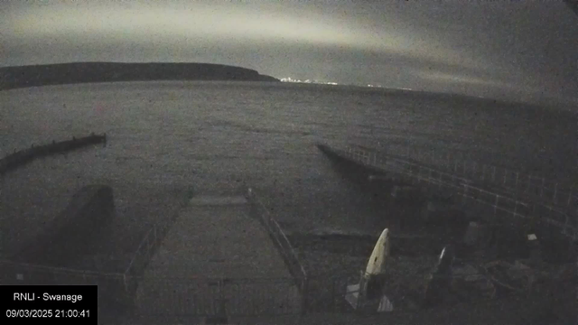 A dark seaside scene at night with a gently rippling ocean. In the foreground, a wooden pier extends into the water, featuring a small boat on it. To the right, there are white and dark objects, possibly equipment or kayaks, partially visible. The background shows a distant shoreline with faint lights indicating a town or a city. The sky has a cloudy appearance, contributing to the overall dim lighting of the scene. The image is timestamped "09/03/2025 21:00:41."