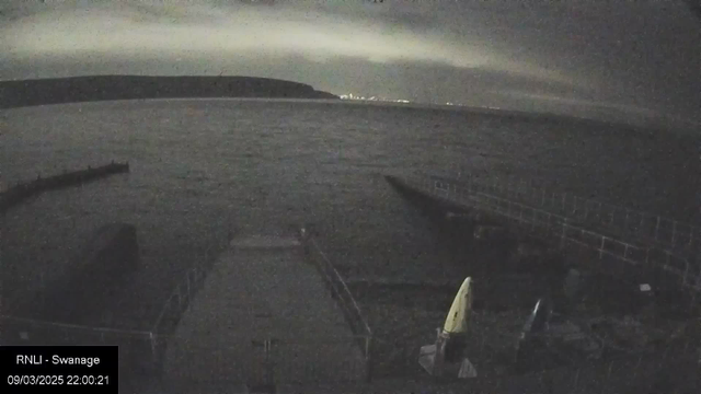 A dimly lit webcam image showing a calm body of water with faint ripples. In the foreground, a narrow pier extends into the water, leading off to the right. There are two kayak-like boats on the pier, one upright with a yellow cover and another lying on its side. In the background, a dark silhouette of a hillside can be seen against the night sky, which is slightly illuminated, hinting at distant lights. The image has a grainy quality, indicating a low-light setting.