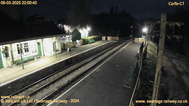 A dimly lit railway station platform at night. The platform features a stone building with large windows, a green bench, and a sign indicating "WAY OUT." On the right side, there is a wooden fence with a couple of green benches positioned along the platform. The railway tracks run parallel to the platform, and the scene is illuminated by a few light sources, creating a tranquil atmosphere.