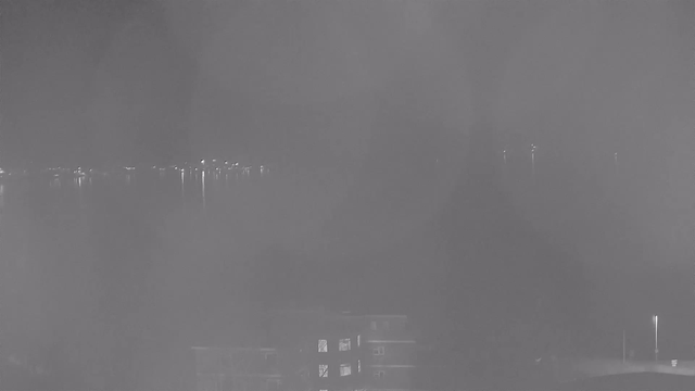 A black-and-white webcam image showing a foggy scene over the water at night. In the distance, faint lights are visible reflecting on the surface of the water. In the foreground, there are blurred outlines of buildings, with some light coming from windows. The overall atmosphere appears hazy and dim.