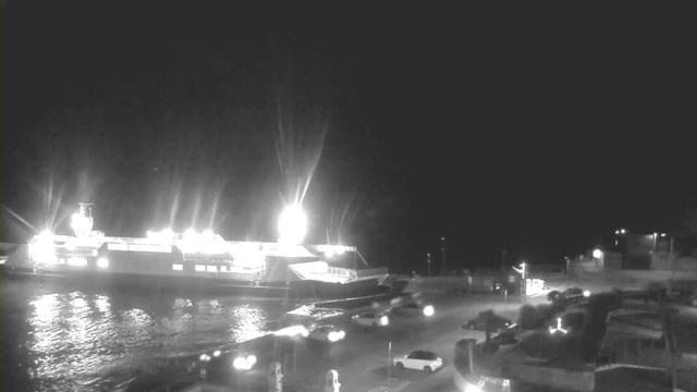 A nighttime scene of a harbor with a large illuminated ferry docked on the left side. The ferry has bright lights shining from its upper deck, reflecting on the water. In the foreground, there are several parked cars and some streetlights illuminating the area. The background is dark with minimal visible structures and faint lights in the distance.