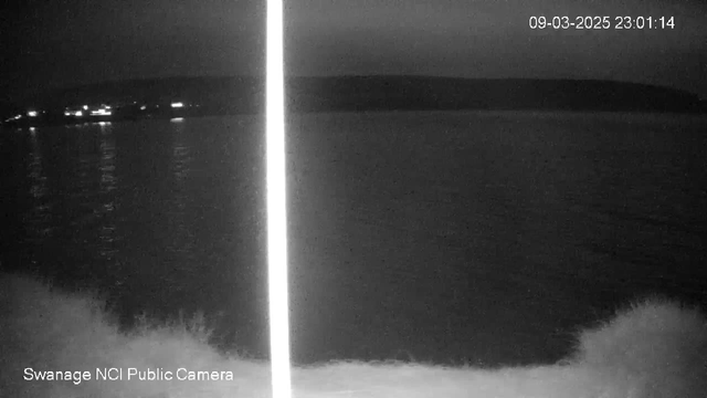 A dark scene captured by a webcam showing a calm body of water. In the background, faint lights from distant structures reflect on the water's surface. The upper half of the image is mostly dark, indicating nighttime, while the lower part shows the water close to the camera. A light vertical line stretches from the top to the bottom of the image. In the bottom left corner, there is a label indicating "Swanage NCI Public Camera." The current timestamp is displayed in the top right corner.