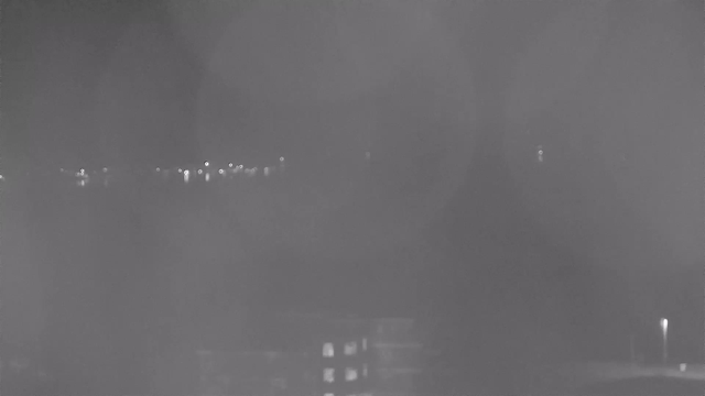 A nighttime webcam view showing a blurred image of distant lights, likely from buildings or street lamps, with a dark, indistinct foreground and background. The scene has a greyish tint and lacks clear details due to low light conditions.