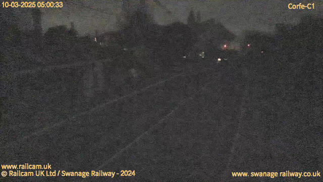 The image depicts a dark scene from a webcam showing a railway area at dawn. The visibility is low, with silhouettes of trees and buildings in the background. There are faint red lights and a shadowy track visible in the foreground, indicating the presence of train infrastructure. The timestamp suggests it is early morning. Overall, the scene conveys a quiet, dimly lit environment with minimal illumination.