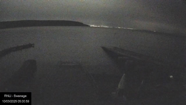 A dimly lit view of a calm body of water at night, with a faint outline of land in the distance. The water reflects minimal light, creating a peaceful but dark atmosphere. In the foreground, there is a wooden structure or pier. The sky is mostly overcast, contributing to the low visibility. A timestamp in the lower left corner indicates the date and time of the image.