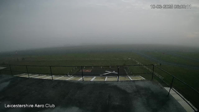 A foggy scene from a webcam overlooking a grassy airfield. The view is obscured by thick mist, making it difficult to see far into the distance. A helipad with a white 'X' marking is visible in the foreground, along with some boundary fencing around the field. The sky is overcast and gray, indicating a cloudy day. The timestamp in the top right corner shows the current date and time.