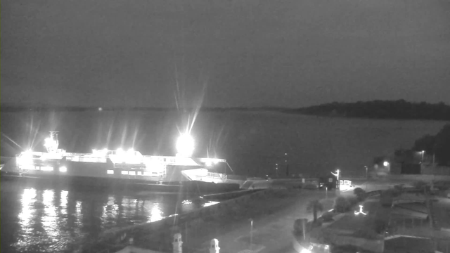 A dark scene featuring a large ferry boat illuminated by bright lights as it sits in calm water. The ferry is docked at a port with a shoreline in the background, partially obscured by darkness. Reflections of the lights shimmer on the water surface, while the surrounding area includes a few buildings and streetlights, suggesting a quiet night atmosphere.