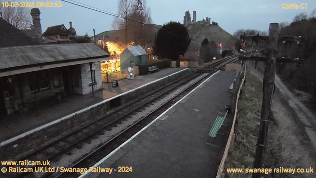 A dimly lit train station scene at dawn, featuring two railway tracks running through the foreground. To the left, a traditional stone building with a sloping roof is partially visible, while just beyond it, a small green wooden shed is illuminated by warm light. On the platform, there are a few green benches and a sign. In the background, a hill rises with ancient castle ruins, and traffic signals stand to the right, showing red lights. The area is bordered by a wooden fence, with a path leading away from the station.