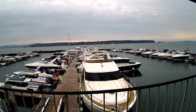 A marina scene featuring numerous boats and yachts moored along a wooden dock. In the foreground, a large white yacht is prominent, with a dark windshield and a canopy. Surrounding it are various smaller boats, primarily in shades of blue and white. The water is calm, and the distant shore is visible in the background, with cloudy skies above and hints of a sunset.