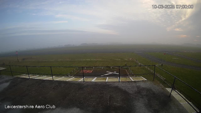 A view from a webcam at Leicestershire Aero Club, showing a grassy airfield shrouded in fog. In the foreground, there's a flat rooftop with a railing and a helipad marked with an 'X'. The horizon features low-lying hills and a cloudy sky with some coloration from the rising sun, indicating early morning. The scene appears serene and quiet, with minimal visibility due to the fog. The date and time are displayed in the top right corner: 10-03-2025, 07:00:00.