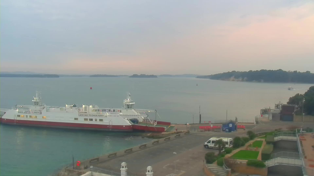 A calm harbor scene features a white and red ferry docked at a pier. The water is a soft blue-green, with gentle ripples reflecting the sky. In the background, several small islands are visible, shrouded in a light mist. The sky is overcast, with hints of pastel colors near the horizon. On the pier, there is a white vehicle parked, and some landscaped areas with grass and plants can be seen in the foreground.