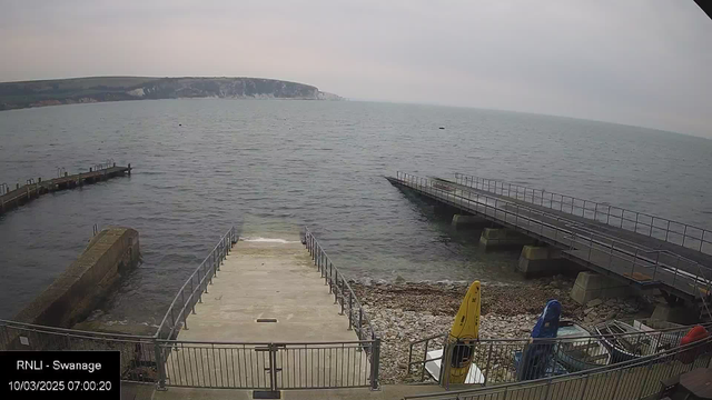 A view from a webcam showing a serene coastal scene. In the foreground, there are stairs leading down to the water, with a railing on either side. To the left, a short pier extends into the sea, and to the right, a longer pier is visible, also extending into the water. Various boats, including some in bright colors (yellow, blue, and red), are parked nearby on the rocky shoreline. The water appears calm, with gentle ripples, and a cloudy sky looms overhead, creating a tranquil atmosphere. In the background, rolling hills and cliffs are visible along the coastline. The image timestamp indicates it was taken on March 10, 2025, at 7:00 AM.