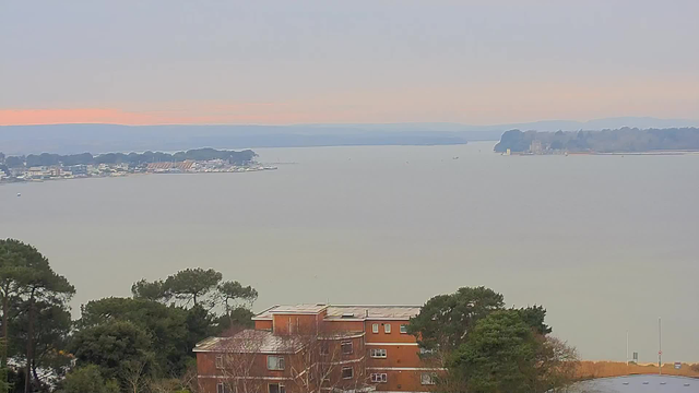 A wide view of a calm body of water, likely a bay or harbor, extending into the distance. The shoreline features several buildings, some of which are low-rise and brick-colored. There are trees in the foreground, and a soft, muted sky with faint hints of orange near the horizon, suggesting early morning or late afternoon. The water appears tranquil, with a few small boats visible in the distance.