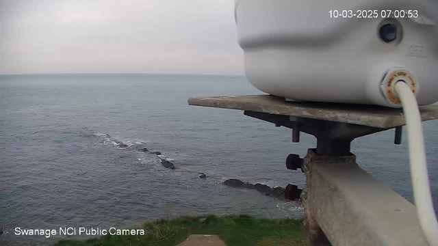 A close-up view of a white device mounted on a metal structure overlooking a calm ocean. The water is a bluish-gray color, with a few dark rocks visible just below the surface. The shoreline is grassy, and the sky above is overcast with soft, muted tones. The timestamp in the corner indicates the date and time.