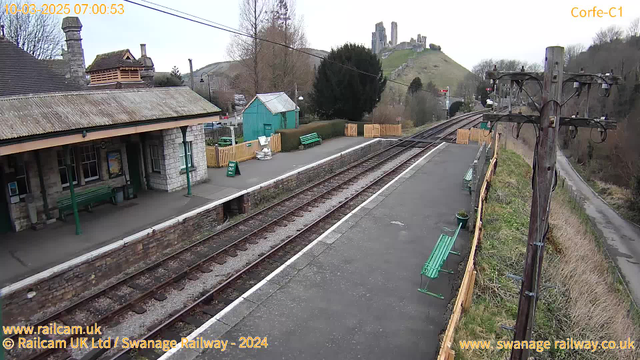 A railway station scene during the day. The platform is lined with several green benches. On the left side, there is a stone building with a peaked roof, and a sign that says "HAY OUT" is visible. To the right, railway tracks stretch into the distance. In the background, a hillside with ruins can be seen, partially obscured by trees. The sky is overcast and gray. A wooden fence borders the platform, and a telegraph pole is positioned on the right side of the image.