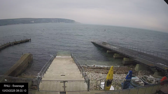 A view of the ocean from a promenade with multiple piers extending into the water. The scene shows a cloudy sky above, with the shoreline visible in the background. There are steps leading down to the water from a concrete platform. To the right, there are brightly colored kayaks, including yellow, blue, and red, resting on a rocky area. The water is calm with some distant waves noticeable. The overall atmosphere appears tranquil and coastal.