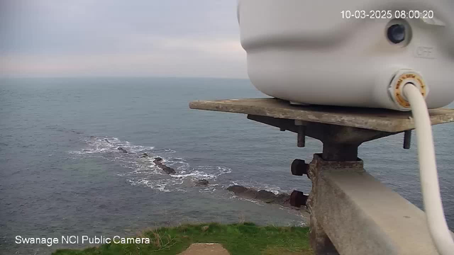 A camera mounted on a pole overlooks a coastal view. The foreground features a sturdy metal support with a white cylindrical object attached. Below, rocky formations are partially visible in the turquoise sea, which stretches toward the horizon under a cloudy sky. The image conveys a calm, serene atmosphere by the water.