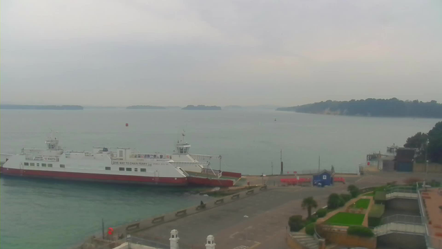 A cloudy scene overlooking a calm body of water with a white and red ferry docked at a pier. The ferry has several visible windows and a ramp extended towards the dock. In the background, there are several small green islands. The shoreline features a road and landscaped areas with plants and palm trees. A small blue building is visible near the dock, along with some construction barriers. The atmosphere is tranquil and somewhat overcast.